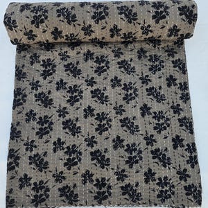 May include: A brown and black floral kantha quilt with a repeating pattern of black flowers on a light brown background. The quilt is rolled up and has a white background.