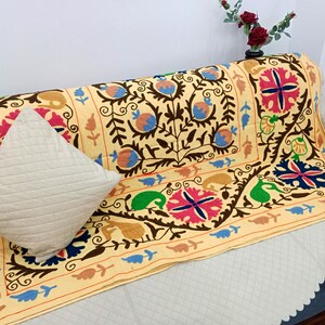 May include: A beige sofa throw with intricate floral embroidery in brown, blue, pink, and green. A matching beige quilted pillow rests on the sofa. The throw features a detailed border design.