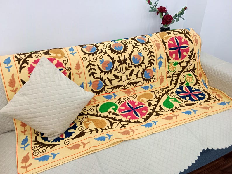 May include: A beige sofa throw with intricate floral embroidery in brown, blue, pink, and green. A matching beige quilted pillow rests on the sofa. The throw features a detailed border design.
