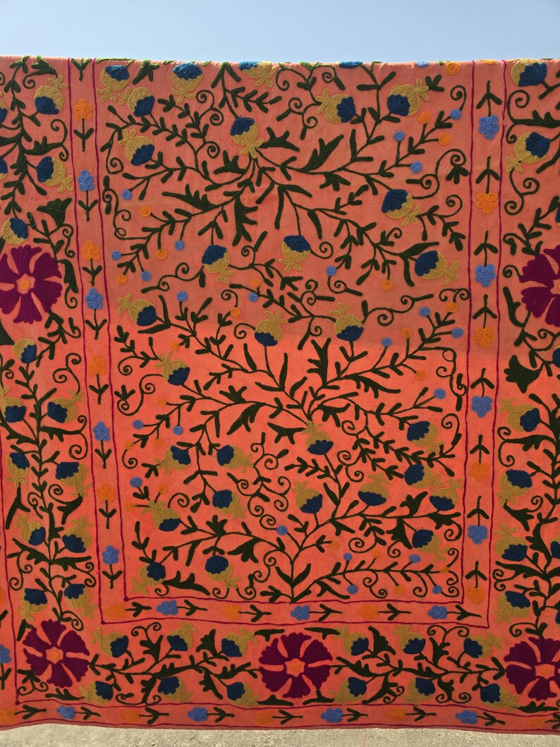 May include: Coral-colored textile with intricate floral embroidery. The design features dark green vines, blue and yellow flowers, and large purple flowers at the corners. The embroidery creates a detailed, textured pattern across the fabric.