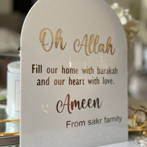 Könnte beinhalten: Ein weißes Acrylschild mit gewölbter Oberseite und rechteckigem Sockel. Goldfarbene Schrift lautet: "Oh Allah, Fülle unser Zuhause mit Barakah und unser Herz mit Liebe. Ameen Von der Familie sakr."
