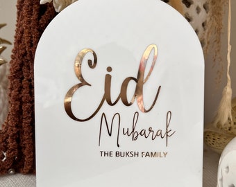 May include: A white, arched sign with gold lettering that reads "Eid Mubarak" and "THE BUKSH FAMILY". The sign is likely made of acrylic or a similar material and is designed for a special occasion.