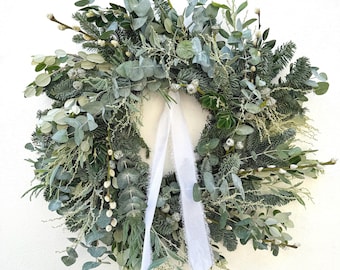 May include: A vibrant green wreath featuring a mix of foliage, such as eucalyptus and pine, with small white buds. A white ribbon is tied at the bottom, creating a natural and elegant appearance. The wreath is a decorative item.