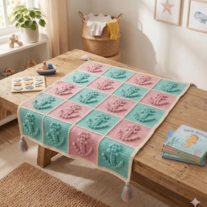 May include: A handmade crochet blanket in a nautical theme. The blanket features a repeating pattern of pink and teal squares, each with a raised anchor design. The blanket has a cream-colored border and tassels at the corners.