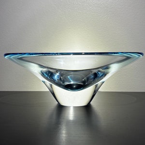 May include: A clear glass bowl with a flared, asymmetrical design. The rim is outlined with a thin band of blue. The bowl sits on a dark surface, with a light background.
