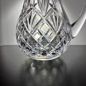 May include: A clear, cut-crystal pitcher with a rounded base and a handle. The pitcher features a diamond and fan pattern, reflecting light and creating a sparkling effect. The pitcher is likely used for serving beverages.