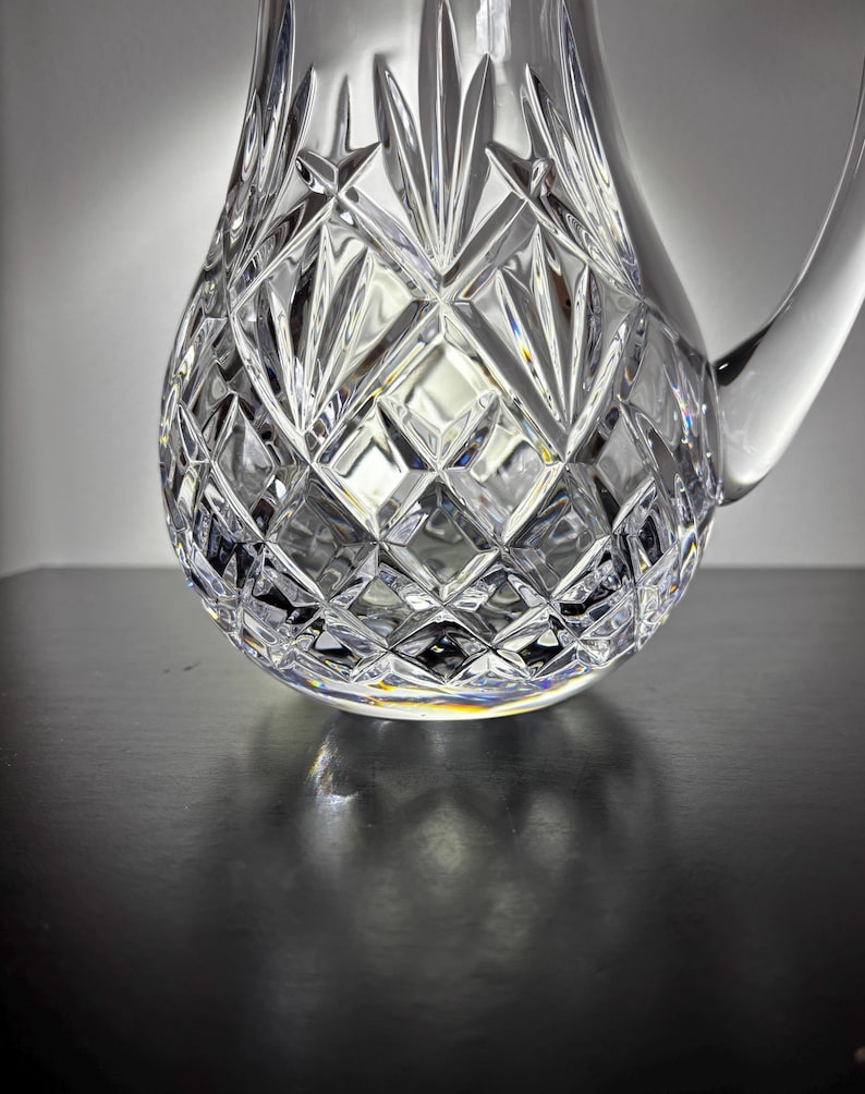 May include: A clear, cut-crystal pitcher with a rounded base and a handle. The pitcher features a diamond and fan pattern, reflecting light and creating a sparkling effect. The pitcher is likely used for serving beverages.