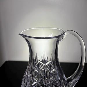 May include: A clear crystal pitcher with a handle. The pitcher has a wide opening and a spout for pouring. The body of the pitcher is decorated with a cut crystal pattern. The pitcher is likely used for serving water or other beverages.