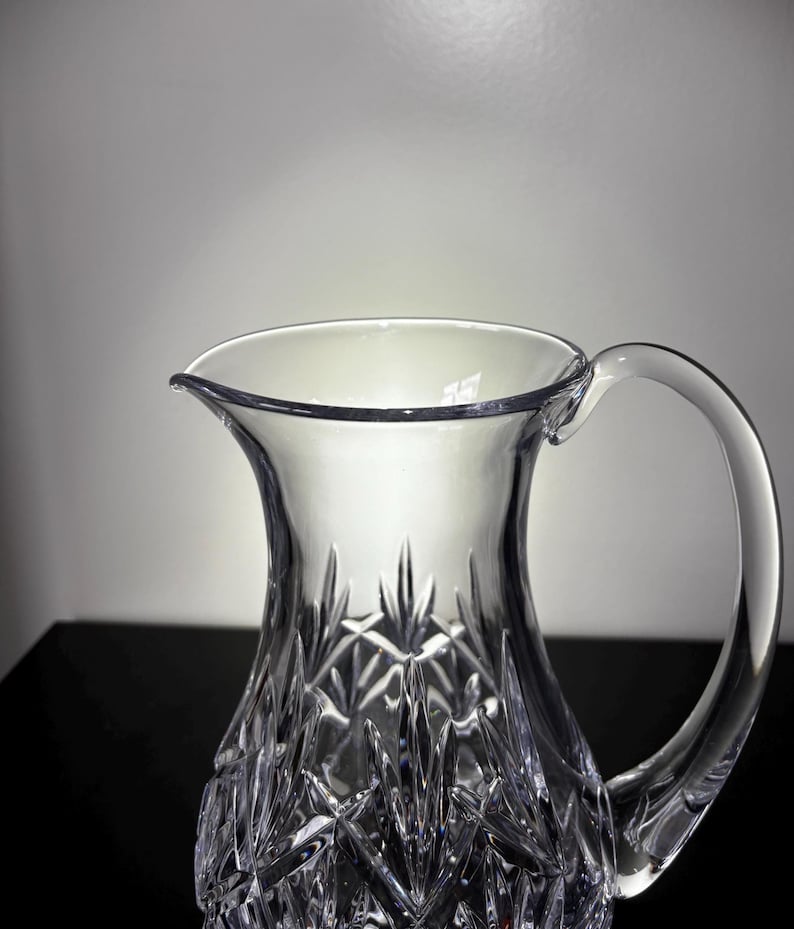 May include: A clear crystal pitcher with a handle. The pitcher has a wide opening and a spout for pouring. The body of the pitcher is decorated with a cut crystal pattern. The pitcher is likely used for serving water or other beverages.