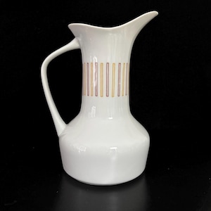 May include: A white ceramic pitcher with a curved handle and a spout. The pitcher features a vertical stripe design in gold and burgundy. The pitcher has a bulbous base and a sleek, modern design.