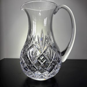 May include: A clear crystal pitcher with a large handle and a spout. The pitcher has a diamond-cut pattern on the lower portion and a smooth, rounded upper section. The handle is curved and attached to the side of the pitcher.