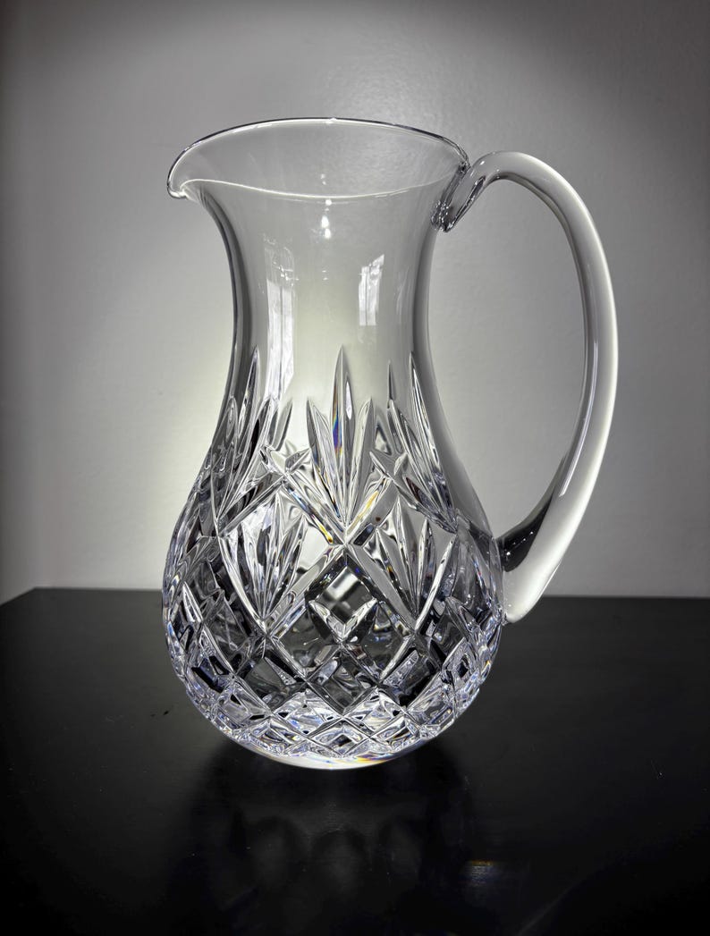 May include: A clear crystal pitcher with a large handle and a spout. The pitcher has a diamond-cut pattern on the lower portion and a smooth, rounded upper section. The handle is curved and attached to the side of the pitcher.