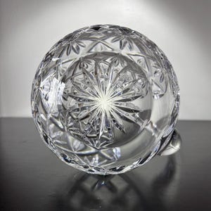 May include: Clear crystal pitcher with intricate diamond and starburst cut patterns. The pitcher has a rounded body and a small handle. The cut glass design reflects light, creating a sparkling effect. A decorative piece for serving beverages.