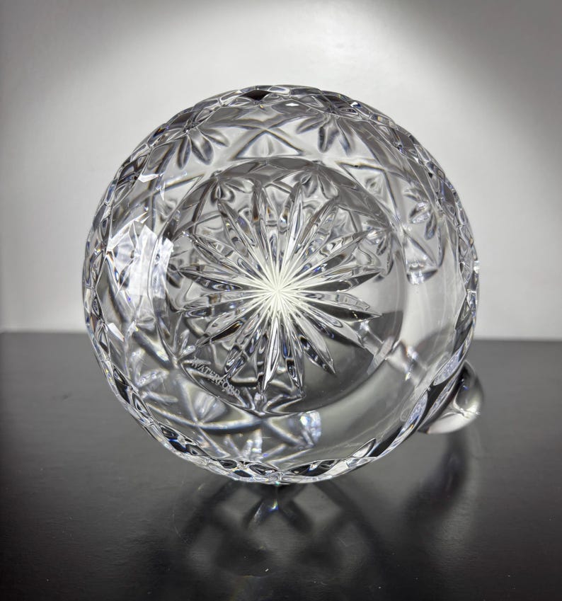May include: Clear crystal pitcher with intricate diamond and starburst cut patterns. The pitcher has a rounded body and a small handle. The cut glass design reflects light, creating a sparkling effect. A decorative piece for serving beverages.