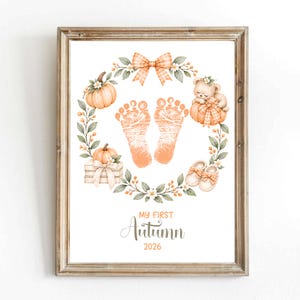May include: A framed print featuring a wreath of orange pumpkins, green leaves, a plaid bow, a teddy bear, and baby shoes surrounding two orange baby footprints. The text reads My First Autumn 2026 in orange and gray script and block lettering.