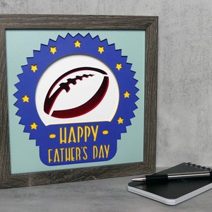 Pode incluir: Um sinal de madeira enquadrado com um design circular azul e amarelo. O design apresenta uma bola de futebol vermelha e branca e o texto "Feliz Dia dos Pais".