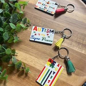 Pode incluir: Três chaveiros com designs coloridos e pingentes. O primeiro chaveiro tem um fundo branco com um unicórnio arco-íris e o texto "Different is Beautiful". O segundo chaveiro tem um fundo branco com o texto "AUTISM is my Super Power" e peças de quebra-cabeça arco-íris. O terceiro chaveiro tem um fundo branco com um design de peça de quebra-cabeça arco-íris e o texto "AUTISM" em um padrão de arco-íris vertical.