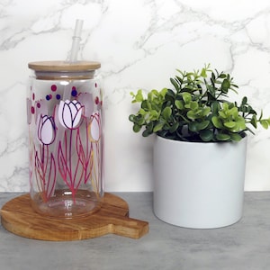 Pode incluir: Uma garrafa de água reutilizável de vidro transparente com tampa de bambu e um canudo. A garrafa tem um design de tulipas rosa, branco e roxo. A garrafa está sobre uma tábua de cortar de madeira.