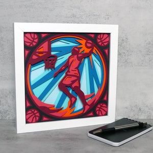 Pode incluir: Uma peça de arte em papel enquadrada em branco que retrata um jogador de basquete em vermelho e azul, fazendo um enterramento. O fundo é um círculo azul e laranja com um contorno preto.