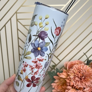 Pode incluir: Um copo de aço inoxidável branco com um design floral com flores rosa, roxas e amarelas. O copo tem uma tampa transparente e um canudo de aço inoxidável.