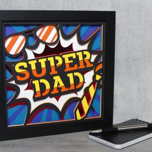 Pode incluir: Uma obra de arte em papel emoldurada e em camadas com as palavras "SUPER DAD" em laranja e amarelo, acentuada com óculos de sol e uma gravata. A obra de arte é colocada sobre um fundo azul e vermelho, com uma moldura preta. Um bloco de notas e uma caneta estão em primeiro plano.