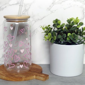 Pode incluir: Uma garrafa de vidro transparente para beber com um padrão de corações rosa e uma tampa de bambu. Um canudo transparente é inserido através da tampa. A garrafa está sentada em uma tábua de cortar de madeira. Uma pequena planta em vaso com folhas verdes está no fundo.