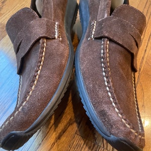 K&ouml;nnte beinhalten: Paar braune Wildleder-Loafer. Diese Slipper haben eine runde Spitze, einen Riemen &uuml;ber dem Oberteil und wei&szlig;e N&auml;hte. Die Sohlen sind dunkelbraun. Geeignet f&uuml;r den Alltag.