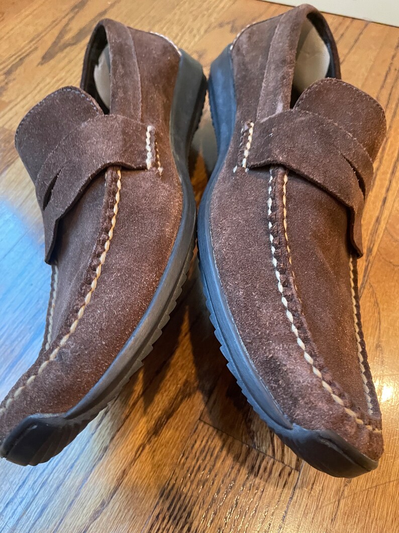 K&ouml;nnte beinhalten: Paar braune Wildleder-Loafer. Diese Slipper haben eine runde Spitze, einen Riemen &uuml;ber dem Oberteil und wei&szlig;e N&auml;hte. Die Sohlen sind dunkelbraun. Geeignet f&uuml;r den Alltag.