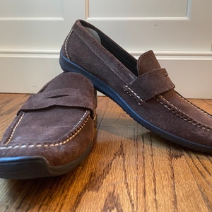 K&ouml;nnte beinhalten: Ein Paar braune Penny-Loafer aus Wildleder mit wei&szlig;en N&auml;hten. Die Schuhe haben ein klassisches Design mit runder Spitze und einem Riemen &uuml;ber dem Spann. Die Sohlen sind schwarz.