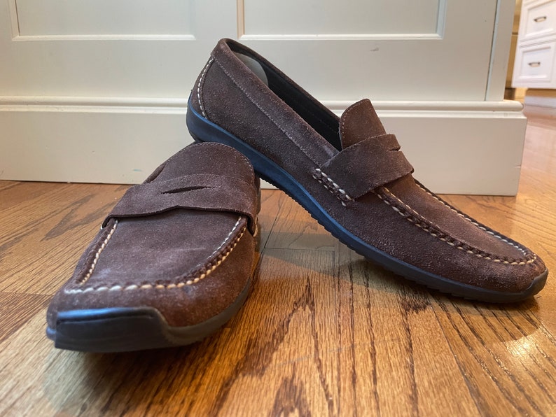 K&ouml;nnte beinhalten: Ein Paar braune Penny-Loafer aus Wildleder mit wei&szlig;en N&auml;hten. Die Schuhe haben ein klassisches Design mit runder Spitze und einem Riemen &uuml;ber dem Spann. Die Sohlen sind schwarz.