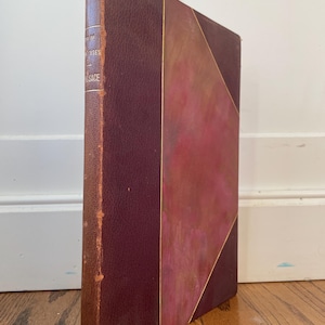 May include: A vintage book with a burgundy leather cover and gold accents. The spine shows the title and author. The cover features a geometric design with a lighter burgundy color. The book is standing upright on a wooden surface.