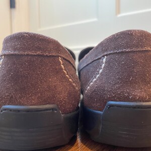 K&ouml;nnte beinhalten: Ein Paar braune Wildleder-Loafer mit schwarzen Sohlen. Die Schuhe haben sichtbare N&auml;hte in einer helleren Farbe entlang der N&auml;hte. Das Bild ist eine Nahaufnahme der Fersen der Schuhe.