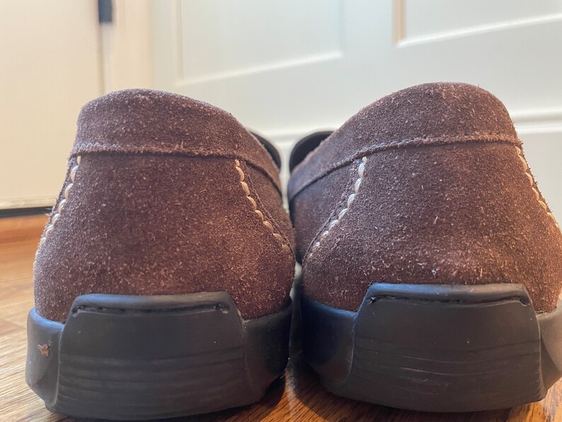 K&ouml;nnte beinhalten: Ein Paar braune Wildleder-Loafer mit schwarzen Sohlen. Die Schuhe haben sichtbare N&auml;hte in einer helleren Farbe entlang der N&auml;hte. Das Bild ist eine Nahaufnahme der Fersen der Schuhe.