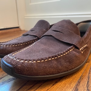 K&ouml;nnte beinhalten: Ein Paar braune Penny-Loafer aus Wildleder mit wei&szlig;en N&auml;hten an den Kanten. Die Schuhe haben ein klassisches Design mit runder Spitze und niedrigem Absatz. Die Loafer werden auf einer Holzoberfl&auml;che pr&auml;sentiert.