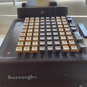 May include: An antique Burroughs adding machine with a dark brown body and beige number keys. The keys are arranged in rows and columns, with numbers from 1 to 7. The word "Burroughs" is printed on the front.