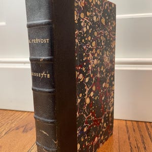 以下が含まれることがあります： 暗い革の背表紙と大理石模様の表紙が付いたアンティーク本。背表紙には金色の文字で「M. Prévost」と「Mussette」のタイトルが記されています。表紙は黒い背景に赤、金、白の大理石模様が施されています。