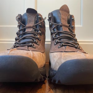 May include: A pair of brown and tan leather boots featuring a rounded rubber toe cap, dark brown laces, and metal eyelets. The boots have a high-top design with a textured rubber sole and a combination of smooth and suede-like leather panels.