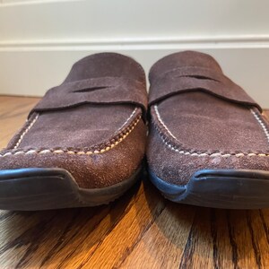 K&ouml;nnte beinhalten: Ein Paar braune Wildleder-Loafer mit Slip-on-Design. Die Schuhe haben eine runde Spitze, einen Riemen &uuml;ber dem Oberteil und wei&szlig;e N&auml;hte an den Kanten. Die Sohlen sind schwarz.