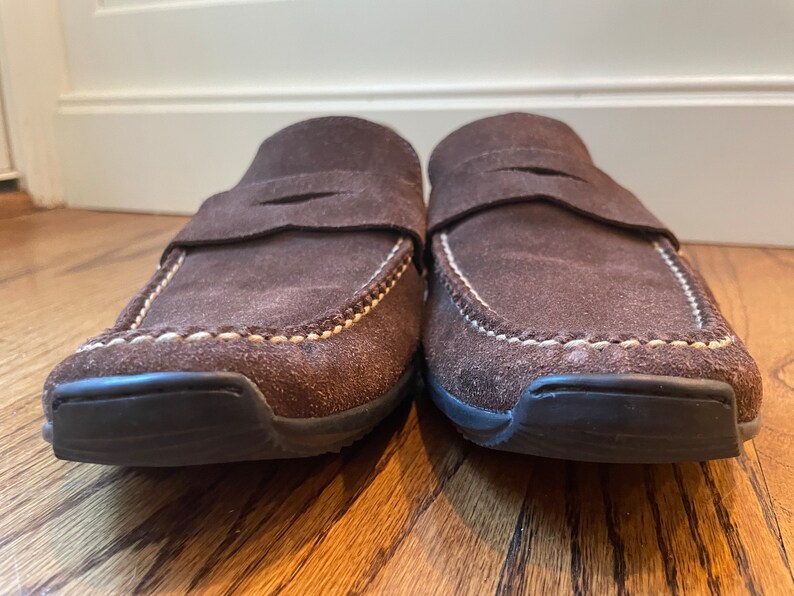 K&ouml;nnte beinhalten: Ein Paar braune Wildleder-Loafer mit Slip-on-Design. Die Schuhe haben eine runde Spitze, einen Riemen &uuml;ber dem Oberteil und wei&szlig;e N&auml;hte an den Kanten. Die Sohlen sind schwarz.