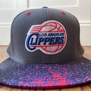 May include: A gray and black baseball cap with a red, white, and blue Los Angeles Clippers logo. The brim of the cap has a pink and blue splatter pattern.
