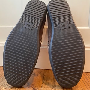 K&ouml;nnte beinhalten: Ein Paar braune Wildlederschuhe, Sohlen nach oben, mit dunkelbraunen Gummisohlen. Die Sohlen haben ein geriffeltes Muster und ein quadratisches Emblem. Die Schuhe stehen auf einem Holzboden.