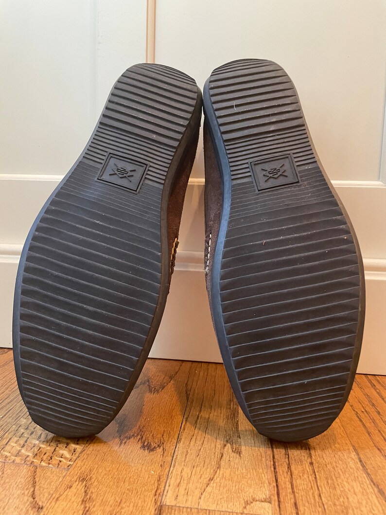 K&ouml;nnte beinhalten: Ein Paar braune Wildlederschuhe, Sohlen nach oben, mit dunkelbraunen Gummisohlen. Die Sohlen haben ein geriffeltes Muster und ein quadratisches Emblem. Die Schuhe stehen auf einem Holzboden.