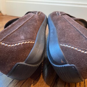 K&ouml;nnte beinhalten: Paar braune Wildleder-Slipper mit Riemen. Die Schuhe haben eine dunkelbraune Sohle und wei&szlig;e N&auml;hte. Diese bequemen Schuhe eignen sich f&uuml;r den Alltag. Die Schuhe sind aus Wildleder gefertigt.