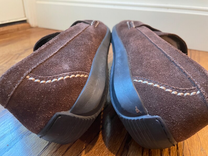 K&ouml;nnte beinhalten: Paar braune Wildleder-Slipper mit Riemen. Die Schuhe haben eine dunkelbraune Sohle und wei&szlig;e N&auml;hte. Diese bequemen Schuhe eignen sich f&uuml;r den Alltag. Die Schuhe sind aus Wildleder gefertigt.
