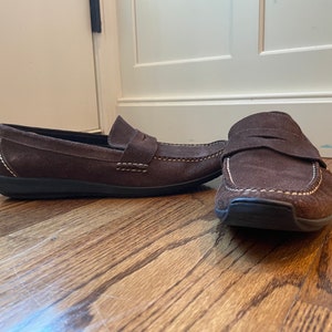 K&ouml;nnte beinhalten: Ein Paar braune Penny-Loafer aus Wildleder mit wei&szlig;en N&auml;hten. Die Schuhe haben ein klassisches Design mit runder Spitze und einem Riemen &uuml;ber dem Spann. Die Sohlen sind schwarz.