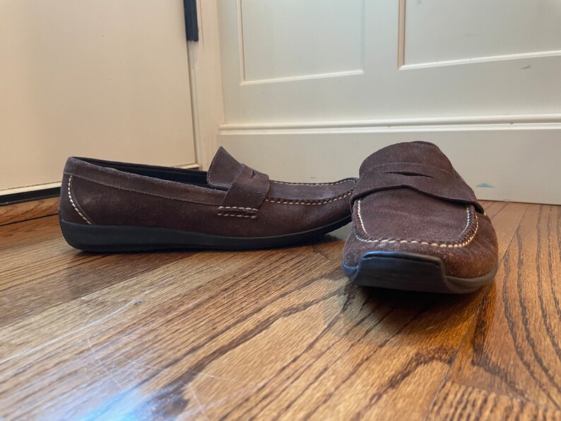 K&ouml;nnte beinhalten: Ein Paar braune Penny-Loafer aus Wildleder mit wei&szlig;en N&auml;hten. Die Schuhe haben ein klassisches Design mit runder Spitze und einem Riemen &uuml;ber dem Spann. Die Sohlen sind schwarz.