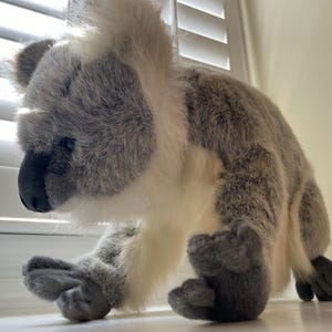 Puede incluir: Un juguete de peluche de koala gris y blanco con una textura suave y peluda. El koala está sentado en una superficie blanca con la cabeza inclinada hacia abajo.