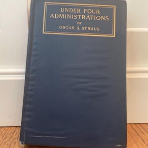 以下が含まれることがあります： オスカー・S・ストラウス著「Under Four Administrations」というタイトルのハードカバー本。タイトルと著者の名前は金色の文字で書かれています。本は白い表面に立てかけられています。