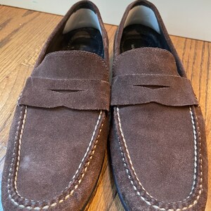 K&ouml;nnte beinhalten: Ein Paar braune Wildleder-Loafer. Diese Schuhe haben ein klassisches Penny-Loafer-Design mit runder Spitze und einem Sattelriemen. Wei&szlig;e N&auml;hte zieren die N&auml;hte. Die Sohlen sind dunkelbraun.