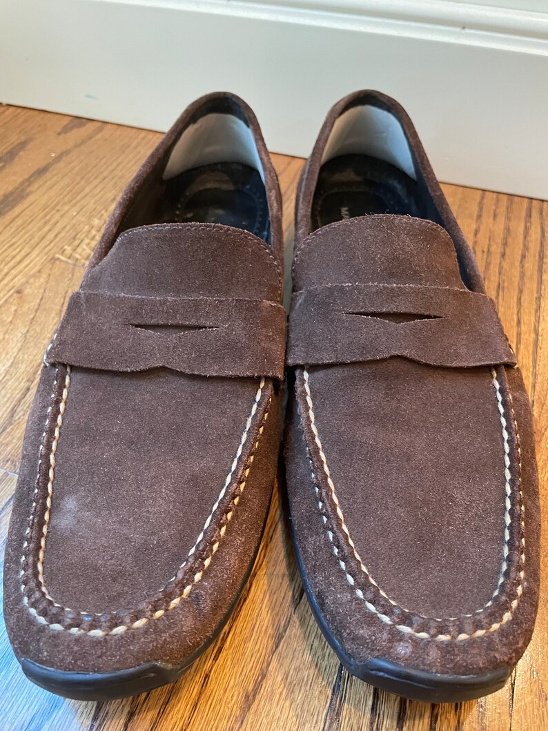 K&ouml;nnte beinhalten: Ein Paar braune Wildleder-Loafer. Diese Schuhe haben ein klassisches Penny-Loafer-Design mit runder Spitze und einem Sattelriemen. Wei&szlig;e N&auml;hte zieren die N&auml;hte. Die Sohlen sind dunkelbraun.